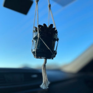 Puede incluir: Una peque&ntilde;a planta suculenta en una maceta azul oscuro cuelga de un macram&eacute; para plantas que est&aacute; sujeto al espejo retrovisor de un coche. La planta est&aacute; enfocada, mientras que el interior del coche est&aacute; desenfocado en el fondo.