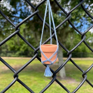 Puede incluir: Una peque&ntilde;a maceta de terracota con un colgador de macram&eacute; azul claro, colgando de una valla de cadena de eslabones negra.