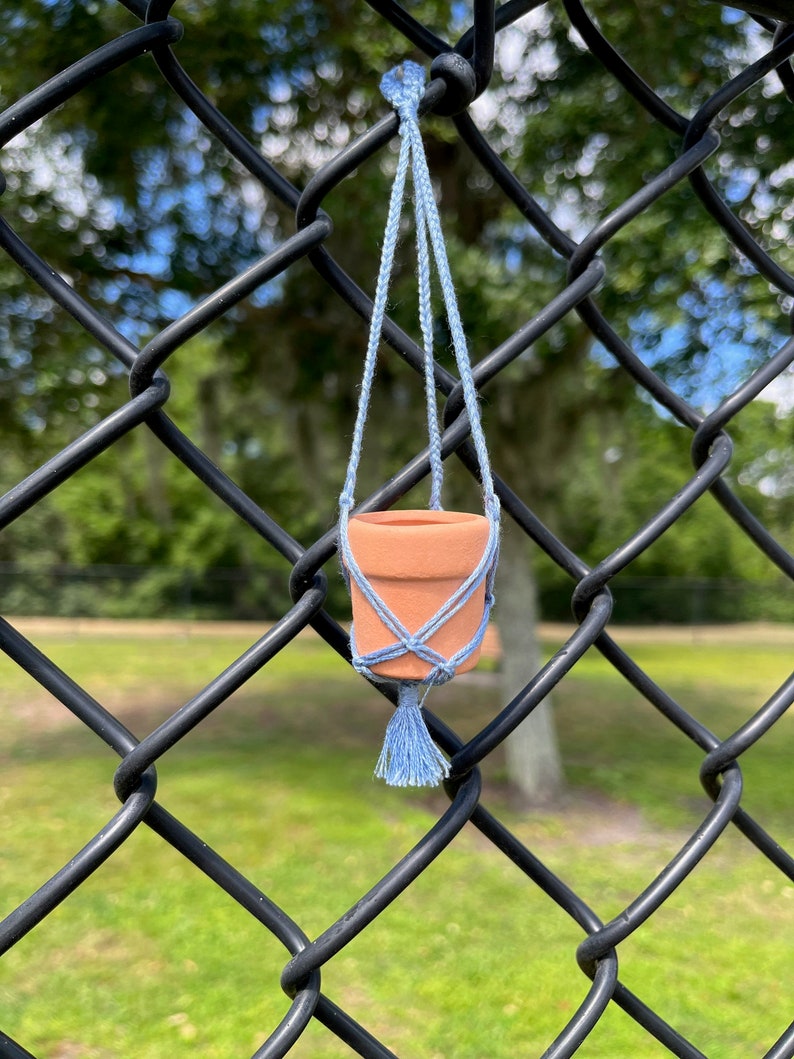 Puede incluir: Una peque&ntilde;a maceta de terracota con un colgador de macram&eacute; azul claro, colgando de una valla de cadena de eslabones negra.