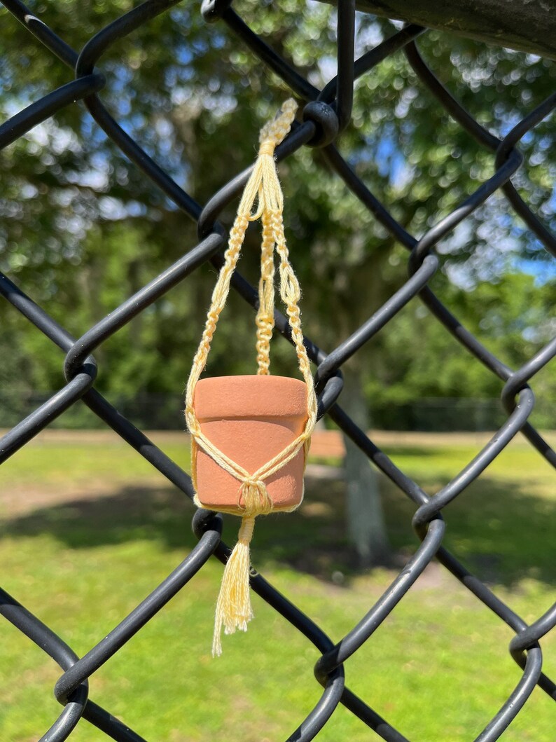 Puede incluir: Una peque&ntilde;a maceta de terracota con un colgador de macram&eacute;, colgando de una valla de cadena de eslabones negra. El macram&eacute; es de color amarillo claro y tiene un borla en la parte inferior.