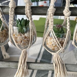 Puede incluir: Tres maceteros de macram&eacute; con peque&ntilde;os frascos de vidrio llenos de guijarros blancos y peque&ntilde;as plantas suculentas verdes. Los maceteros est&aacute;n hechos de cuerda beige natural con borlas.
