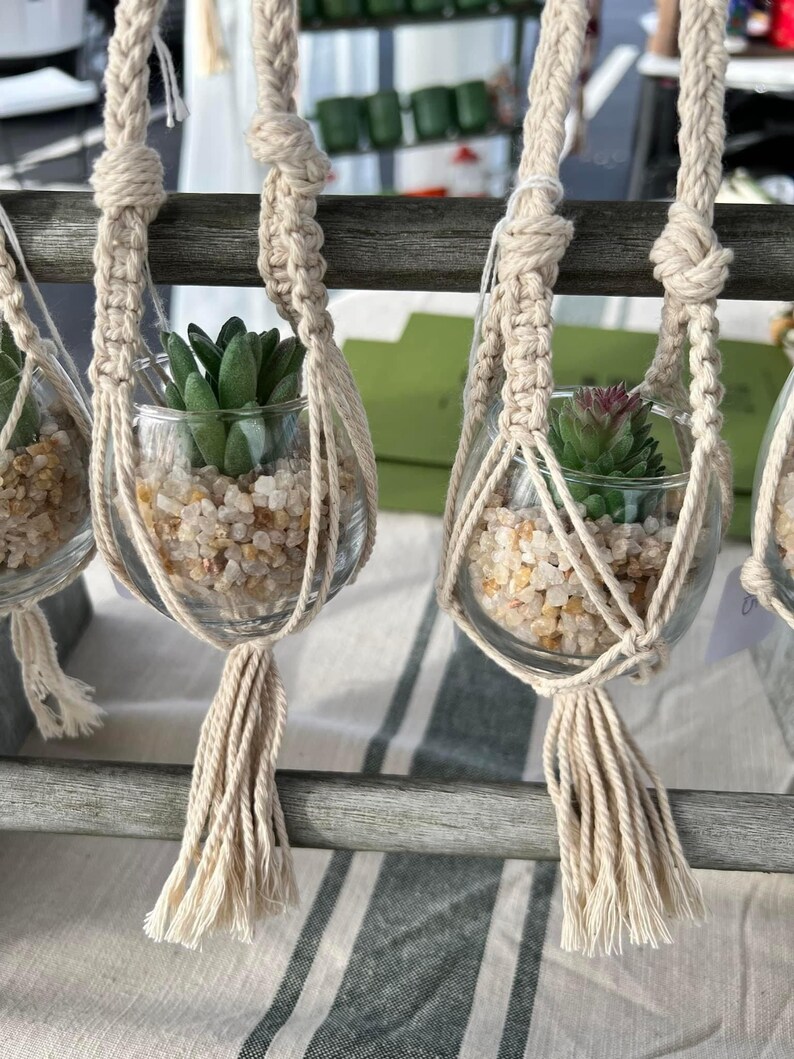 Puede incluir: Tres maceteros de macram&eacute; con peque&ntilde;os frascos de vidrio llenos de guijarros blancos y peque&ntilde;as plantas suculentas verdes. Los maceteros est&aacute;n hechos de cuerda beige natural con borlas.