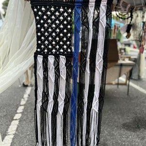 May include: A black and white macrame wall hanging with a blue stripe. The hanging is made with a knotting technique and has long fringe.