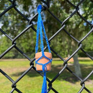 Puede incluir: Una peque&ntilde;a maceta de terracota con un macram&eacute; azul para colgar plantas, colgando de una valla de cadena de eslabones negra.