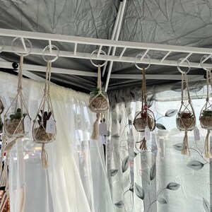 Puede incluir: Un conjunto de ocho terrarios de vidrio colgantes con macram&eacute; para colgar plantas. Cada terrario contiene una peque&ntilde;a planta suculenta y piedras blancas. Los terrarios est&aacute;n colgados de una rejilla de metal blanco.