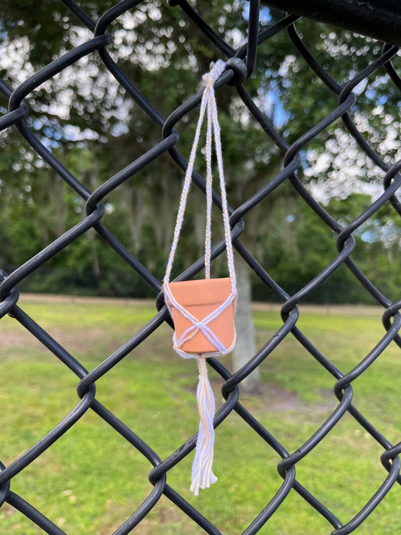 Puede incluir: Una peque&ntilde;a maceta de terracota colgando de una valla de cadena. La maceta est&aacute; suspendida por un macram&eacute; para plantas con un borla blanco y beige.
