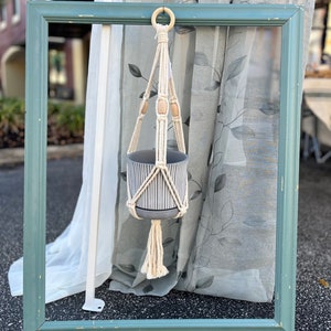 May include: A white macrame plant hanger with wooden beads hangs from a green picture frame. A gray and white striped planter is suspended from the hanger.