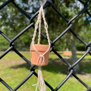 Pode incluir: Um pequeno vaso de terracota com um suporte de macramé, pendurado em uma cerca de arame farpado preta. O vaso está vazio e o suporte é feito de corda bege natural.