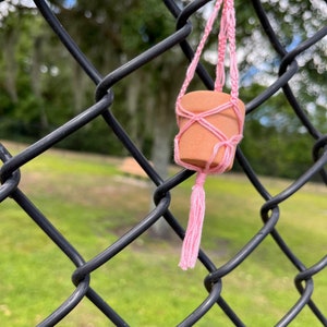 Puede incluir: Una peque&ntilde;a maceta de terracota con un colgador de macram&eacute; rosa colgando de una valla de cadena de eslabones negra.