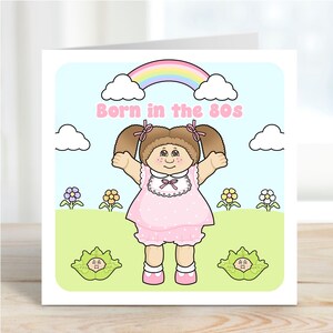 May include: A square greeting card with a cartoon illustration of a doll in a pink dress and bloomers, with brown pigtails. The text "Born in the 80s" is above the doll, with a rainbow and clouds in the background. Flowers and cabbage patch kids are also present.