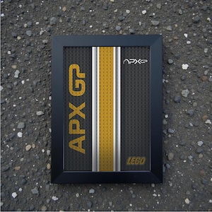 May include: Framed artwork featuring a design with yellow, white, and black stripes, and the text "APX GP" in yellow. The word "LEGO" is also visible. The frame is black.