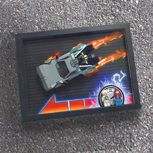 May include: Framed artwork featuring a Lego DeLorean time machine with flames, a red and orange arrow, and Lego figures. The artwork is set against a black background and framed in a black frame.