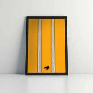 May include: Framed art print featuring a vibrant yellow background with two vertical tire track patterns in white. A black logo is at the bottom. The artwork is encased in a black frame, suitable for wall decor.