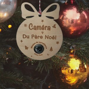 Peut inclure: Ornement de Noël en bois en forme de boule avec un nœud en haut. L'ornement porte l'inscription "Caméra Du Père Noël" et un petit objectif de caméra. Il est suspendu à un sapin de Noël orné de boules rouges et dorées.