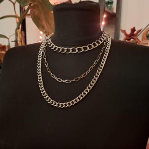 Może przedstawiać: Srebrny naszyjnik z łańcucha o trzech warstwach z trzema różnymi stylami łańcucha. Naszyjnik jest prezentowany na czarnym manekinie.
