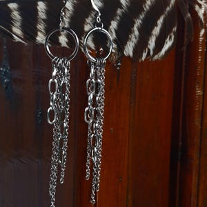 Pode incluir: Um par de brincos de corrente prateada com várias fitas de corrente e um aro circular no topo.