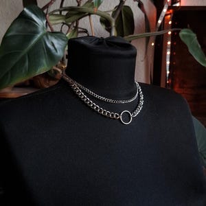 Może przedstawiać: Srebrny naszyjnik z łańcuszka o dwuwarstwowym wzorze i małą okrągłą zawieszką. Naszyjnik jest prezentowany na czarnym golfie, z zieloną rośliną w tle.