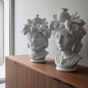Op de afbeelding: Twee witte keramische bustes van klassieke figuren, een man en een vrouw, versierd met fruit- en bladkransen. De bustes staan op een donkerhouten kast met een minimalistisch design, tegen een neutrale muur.