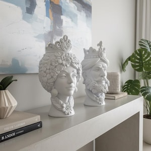Può includere: Due sculture di teste in ceramica bianca, una di una donna con corona floreale e l'altra di un uomo con turbante e barba. Sono esposte su una consolle grigio chiaro, con un dipinto sullo sfondo.