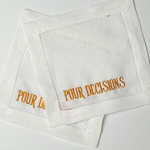 May include: Two white linen coasters with embroidered text in a gold color. The text on the coasters reads "POUR DECISIONS". The coasters have a stitched border.
