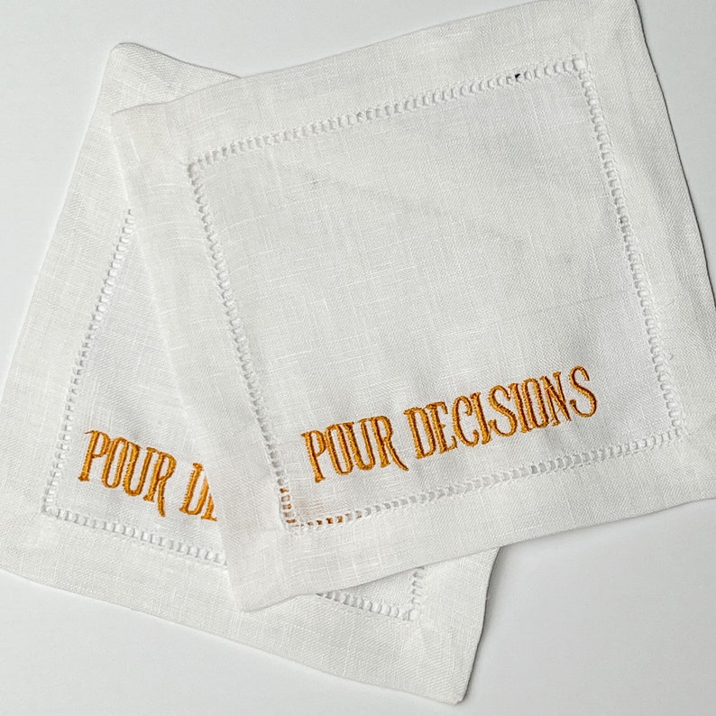 May include: Two white linen coasters with embroidered text in a gold color. The text on the coasters reads "POUR DECISIONS". The coasters have a stitched border.