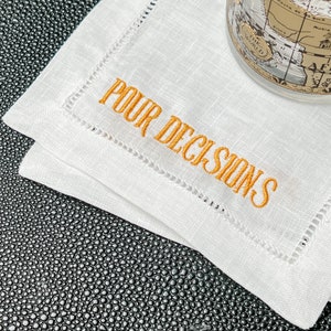 May include: White linen cocktail napkins with orange embroidered text that reads "POUR DECISIONS". The napkins have a stitched border and are stacked on a textured black surface. A globe-shaped glass is partially visible.