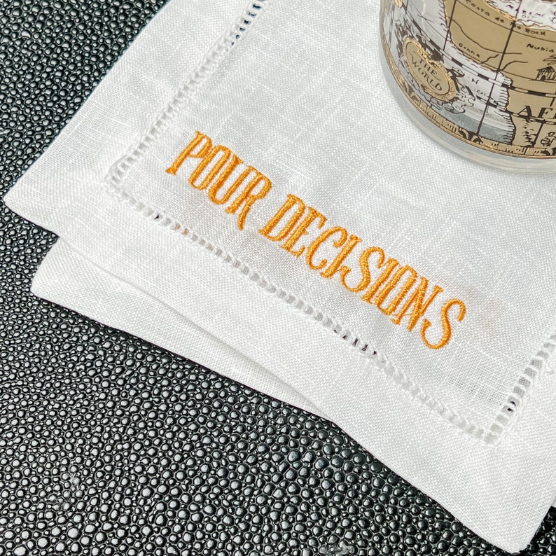 May include: White linen cocktail napkins with orange embroidered text that reads "POUR DECISIONS". The napkins have a stitched border and are stacked on a textured black surface. A globe-shaped glass is partially visible.