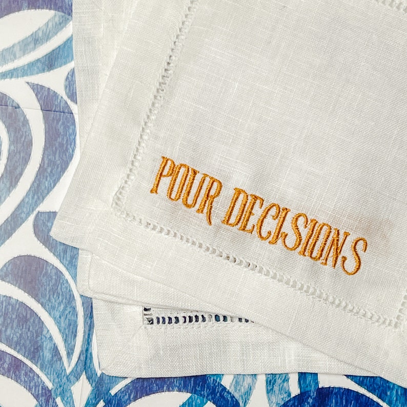 May include: White linen cocktail napkins with embroidered text in gold that reads "POUR DECISIONS". The napkins have a decorative stitched edge. The napkins are stacked on a blue and white patterned surface.