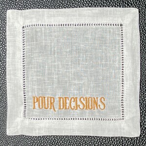 May include: Square white linen coaster with embroidered text "POUR DECISIONS" in gold. The coaster has a stitched border with a decorative pattern. The background is a textured, light gray color.