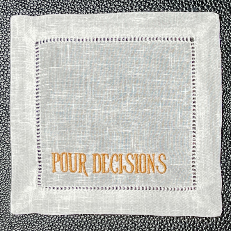 May include: Square white linen coaster with embroidered text "POUR DECISIONS" in gold. The coaster has a stitched border with a decorative pattern. The background is a textured, light gray color.