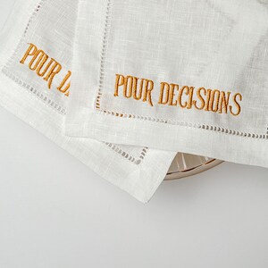 May include: Two white linen cocktail napkins with gold embroidered text that reads "POUR DECISIONS". The napkins have a decorative stitched border and are placed on a light-colored surface.