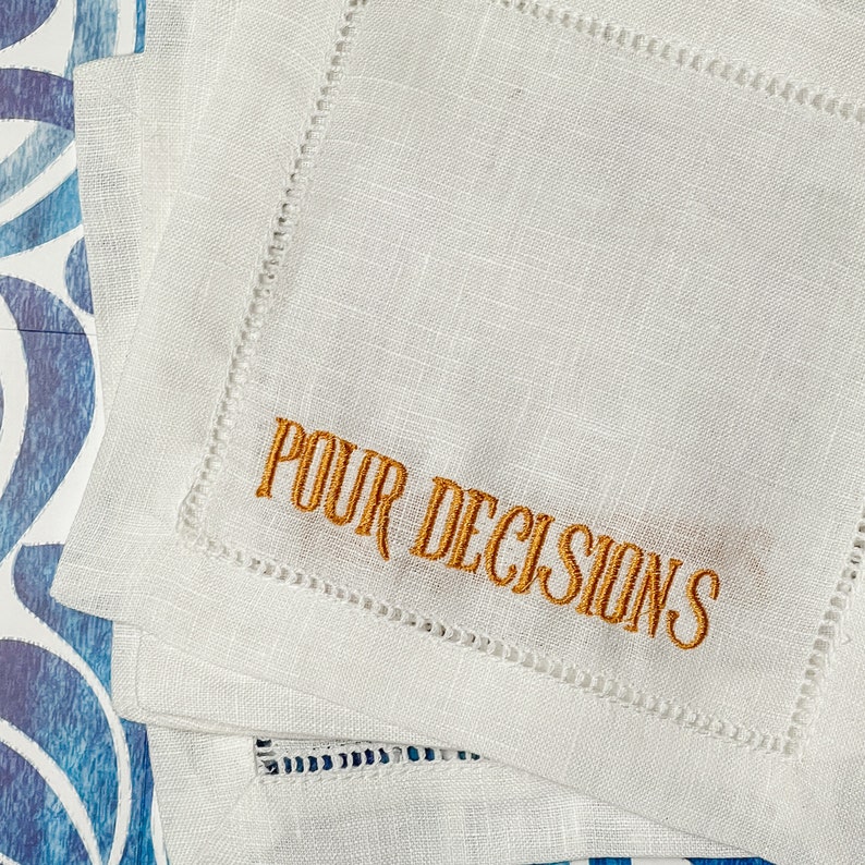 May include: White linen cocktail napkins with embroidered text "POUR DECISIONS" in gold thread. The napkins have a stitched border and are stacked on a blue and white patterned surface. Ideal for a bar or home entertaining.