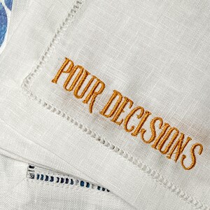 May include: White linen cocktail napkins with the embroidered phrase "POUR DECISIONS" in gold thread. The napkins have a decorative stitched border. The image shows a close-up of the napkins.