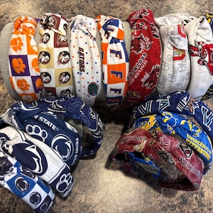Könnte beinhalten: Eine Sammlung von Stoffstirnbändern in verschiedenen Farben und Mustern. Einige Stirnbänder zeigen Logos und Farben von College-Teams, darunter Orange, Blau, Rot und Weiß. Die Stirnbänder sind oben geknotet und scheinen aus weichem Stoff zu sein.