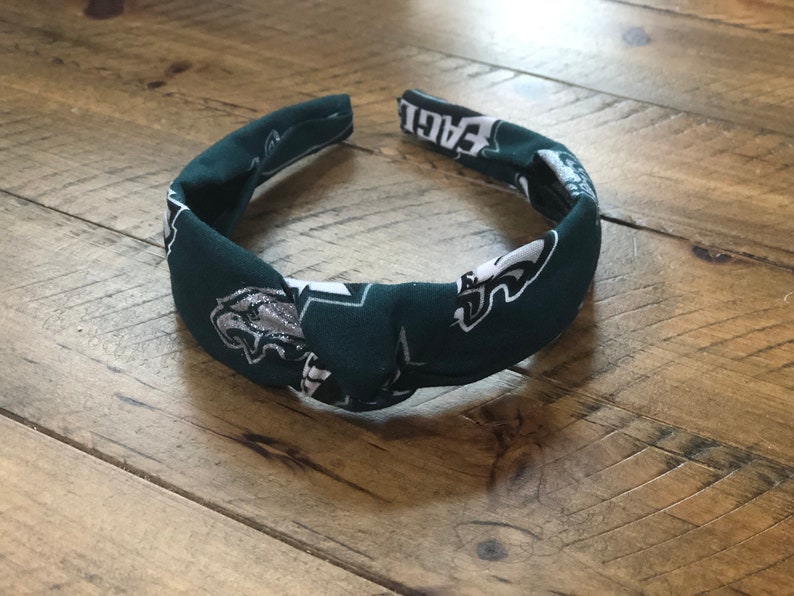 May include: A dark green headband with a knotted design. The fabric features a repeating pattern of the Philadelphia Eagles logo in silver and white. The word "EAGLES" is printed on the headband.