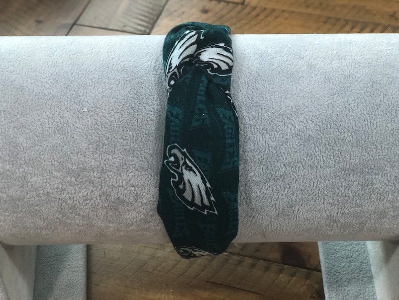 May include: A dark green headband with a repeating pattern of white Philadelphia Eagles logos and text. The headband is made of fabric and is folded over a gray, textured surface.