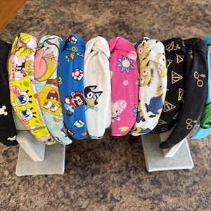 May include: A collection of fabric headbands with various cartoon character prints, including Mickey Mouse, Spongebob, and Harry Potter symbols. The headbands are displayed on a gray stand, showcasing a variety of colors and patterns.