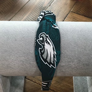 May include: A fabric headband featuring the Philadelphia Eagles logo in white, gray, and black on a dark green background. The headband is wrapped around a gray cylindrical object.