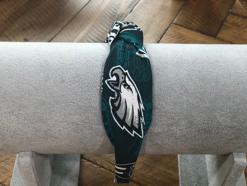 May include: A fabric headband featuring the Philadelphia Eagles logo in white, gray, and black on a dark green background. The headband is wrapped around a gray cylindrical object.