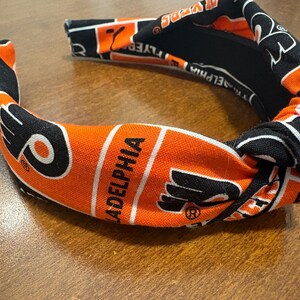 May include: A fabric headband in orange, black, and white, featuring the word "Philadelphia" and a logo. The headband has a knotted design and is likely for sports fans.