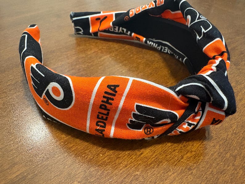 May include: A fabric headband in orange, black, and white, featuring the word "Philadelphia" and a logo. The headband has a knotted design and is likely for sports fans.