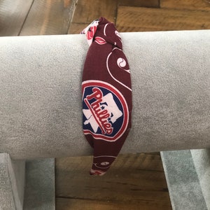 May include: A maroon headband with a repeating pattern and the Philadelphia Phillies logo. The logo is a white baseball bat and the word "Phillies" in red script, set against a blue background.