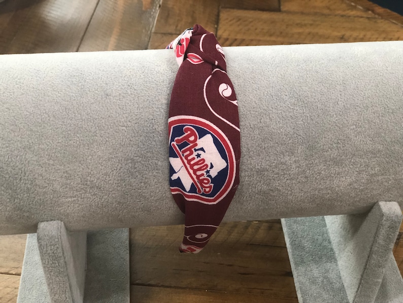 May include: A maroon headband with a repeating pattern and the Philadelphia Phillies logo. The logo is a white baseball bat and the word "Phillies" in red script, set against a blue background.