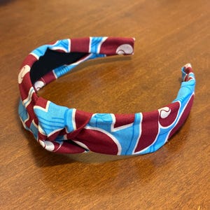 May include: A fabric headband with a knotted design. The headband features a pattern of light blue and white shapes on a maroon background. The interior is black.