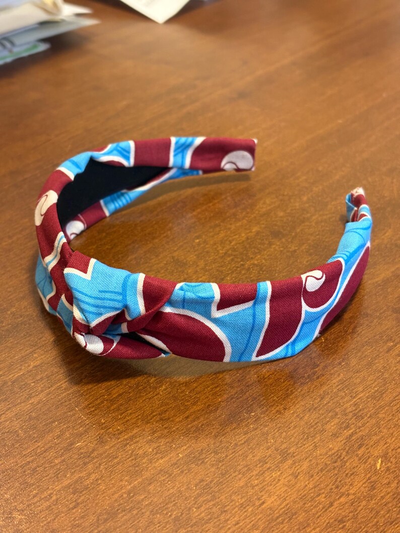 May include: A fabric headband with a knotted design. The headband features a pattern of light blue and white shapes on a maroon background. The interior is black.