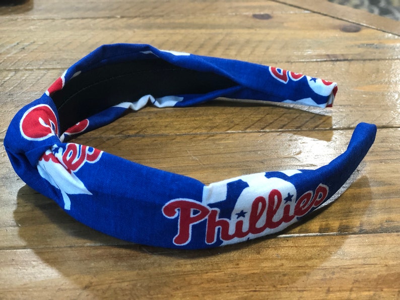 May include: A blue headband featuring the Philadelphia Phillies logo in red and white. The headband is made of fabric and has a knotted design. The Phillies logo is repeated around the headband.