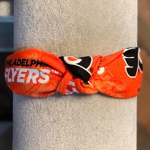 May include: Orange and black Philadelphia Flyers headband. The headband features the team's name in white block letters and the Flyers logo. The headband is knotted and appears to be made of a soft, flexible material.