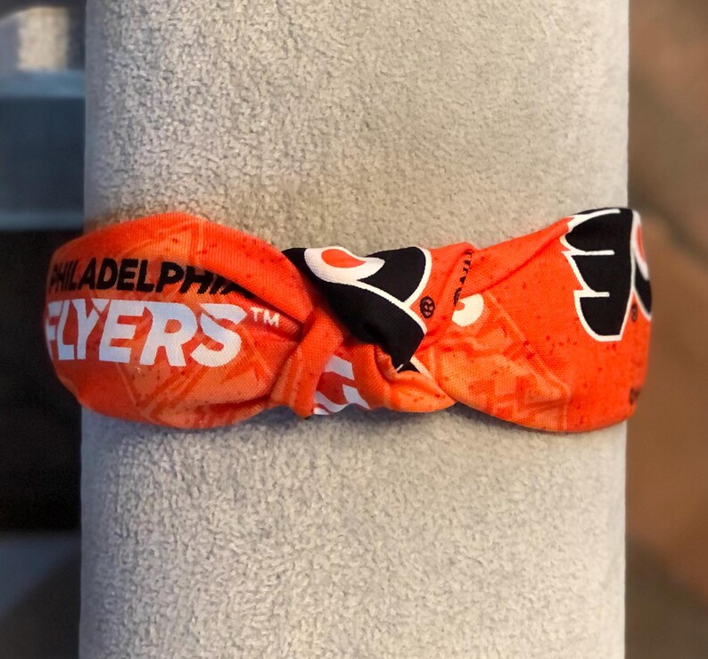 May include: Orange and black Philadelphia Flyers headband. The headband features the team's name in white block letters and the Flyers logo. The headband is knotted and appears to be made of a soft, flexible material.