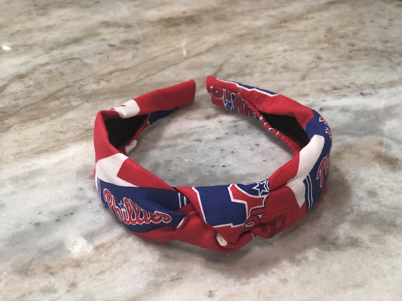 May include: A red, white, and blue fabric headband with the Philadelphia Phillies logo. The headband is knotted at the top and features the team's name and logo in white script on a blue background.