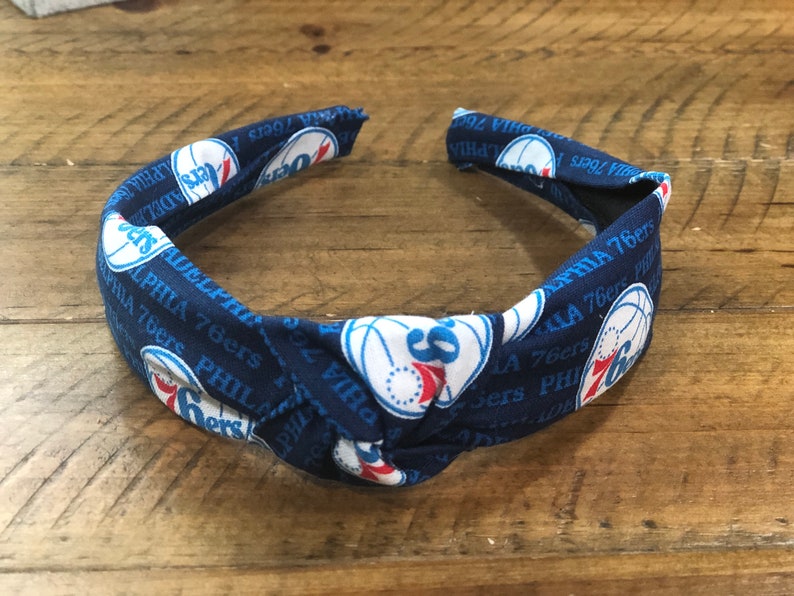 May include: A navy blue headband featuring the Philadelphia 76ers logo and text. The fabric design includes the team's logo, a basketball, and the words "PHILADELPHIA 76ers" in white. The headband is knotted at the top.
